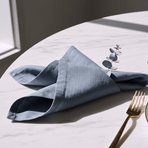 Folded blue napkin with a leaf design on a white tablecloth, accompanied by a gold fork.